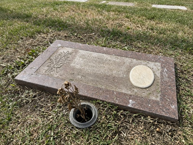 Illustrative example, before: a flat red granite marker with a blank photo plaque, grass creeping over its edges, water stains, and dead stems in the vase